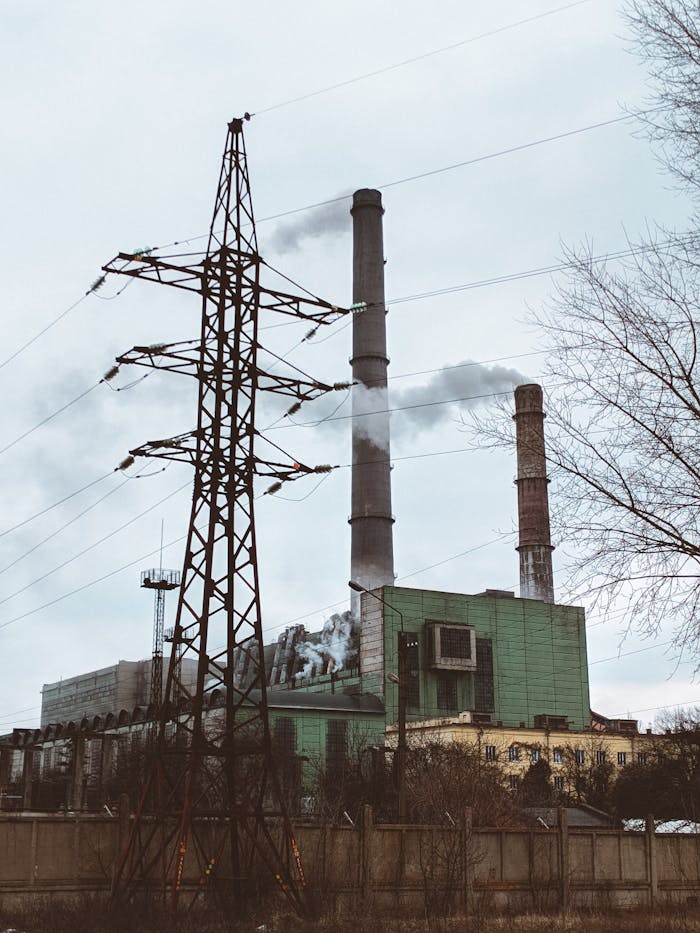A power plant with towering smokestacks emitting smoke beside a high voltage pylon on an overcast day.